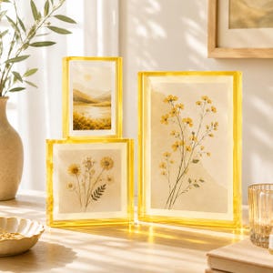 May include: Three rectangular acrylic photo frames with a vibrant yellow tint. The frames display botanical prints of yellow wildflowers and a landscape scene. The frames are clear, thick, and have a polished finish, showcasing the artwork inside.