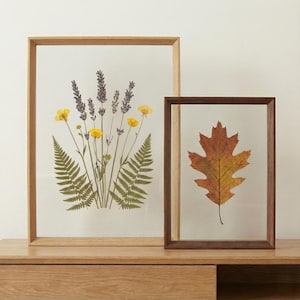 May include: Two framed botanical prints. One features dried lavender, yellow flowers, and ferns in a light wood frame. The other showcases a single autumn leaf in a dark wood frame. Both are displayed on a wooden surface.