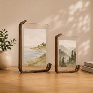 K&ouml;nnte beinhalten: Zwei vertikale Bilderrahmen mit dunkelbraunen Holzsockeln und transparenten Acrylplatten. Der gr&ouml;&szlig;ere Rahmen zeigt eine Aquarell-Landschaft mit einem K&uuml;stenhaus, w&auml;hrend der kleinere Rahmen eine Aquarell-Illustration eines dichten Nadelwaldes enth&auml;lt.