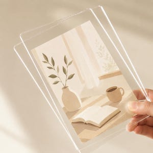 K&ouml;nnte beinhalten: Ein klarer Acrylrahmen mit einem rechteckigen Druck, der eine beigefarbene Illustration einer Vase mit belaubten Zweigen, einer Kaffeetasse und einem aufgeschlagenen Buch auf einem Tisch zeigt. Das Bild verwendet weiche, neutrale Farbt&ouml;ne.
