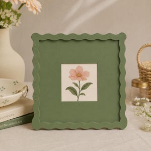 May include: A square picture frame with a matte olive green finish and a scalloped edge. The frame contains a centered square print featuring a botanical illustration of a single pink flower with green leaves on a cream-colored background.