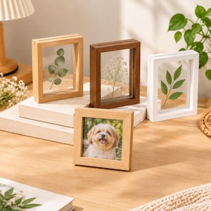 May include: A collection of four rectangular and square picture frames in light wood, dark wood, and white finishes. Three frames feature clear glass panels displaying pressed green leaves and white flowers, while one frame holds a photo of a dog.
