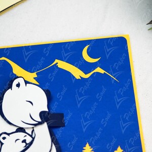 May include: A blue and yellow paper card with a pop-up design of two polar bears hugging. The larger bear is holding a red heart. The background features a yellow crescent moon and yellow mountains against a blue sky.