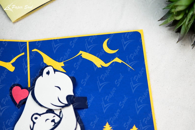 May include: A blue and yellow paper card with a pop-up design of two polar bears hugging. The larger bear is holding a red heart. The background features a yellow crescent moon and yellow mountains against a blue sky.