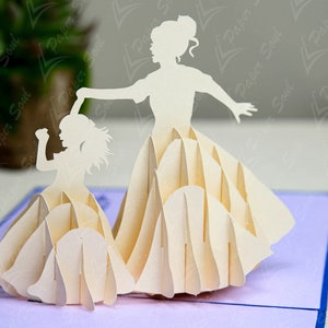 May include: A white paper pop-up card featuring two figures of women in dresses. The figures are layered and appear to be dancing. The card is on a purple background.