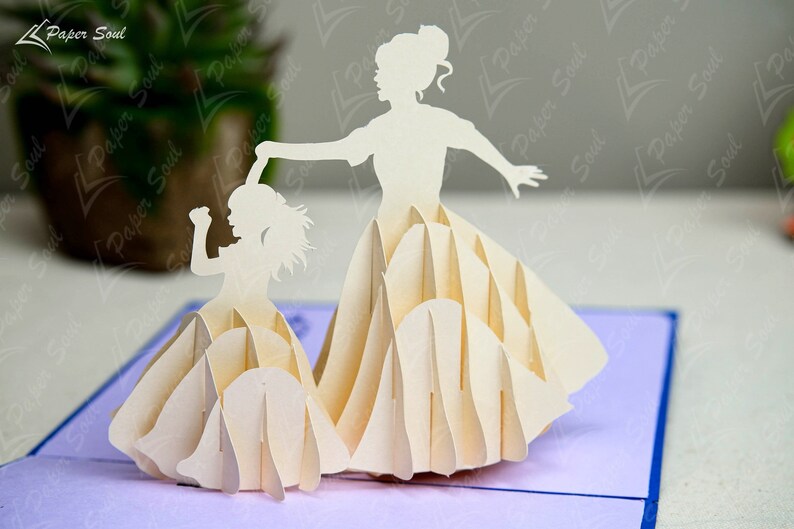 May include: A white paper pop-up card featuring two figures of women in dresses. The figures are layered and appear to be dancing. The card is on a purple background.