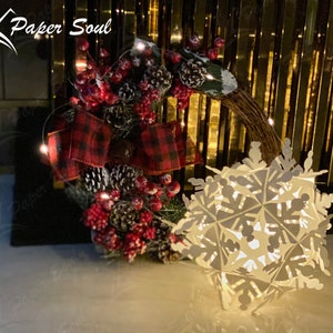 Peut inclure: Une d&eacute;coration de flocon de neige en papier blanc avec une guirlande de lumi&egrave;res blanches enroul&eacute;e autour d'un n&oelig;ud en plaid rouge et noir et une couronne de No&euml;l avec des baies rouges et des pommes de pin.