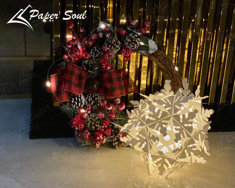 Peut inclure: Une d&eacute;coration de flocon de neige en papier blanc avec une guirlande de lumi&egrave;res blanches enroul&eacute;e autour d'un n&oelig;ud en plaid rouge et noir et une couronne de No&euml;l avec des baies rouges et des pommes de pin.