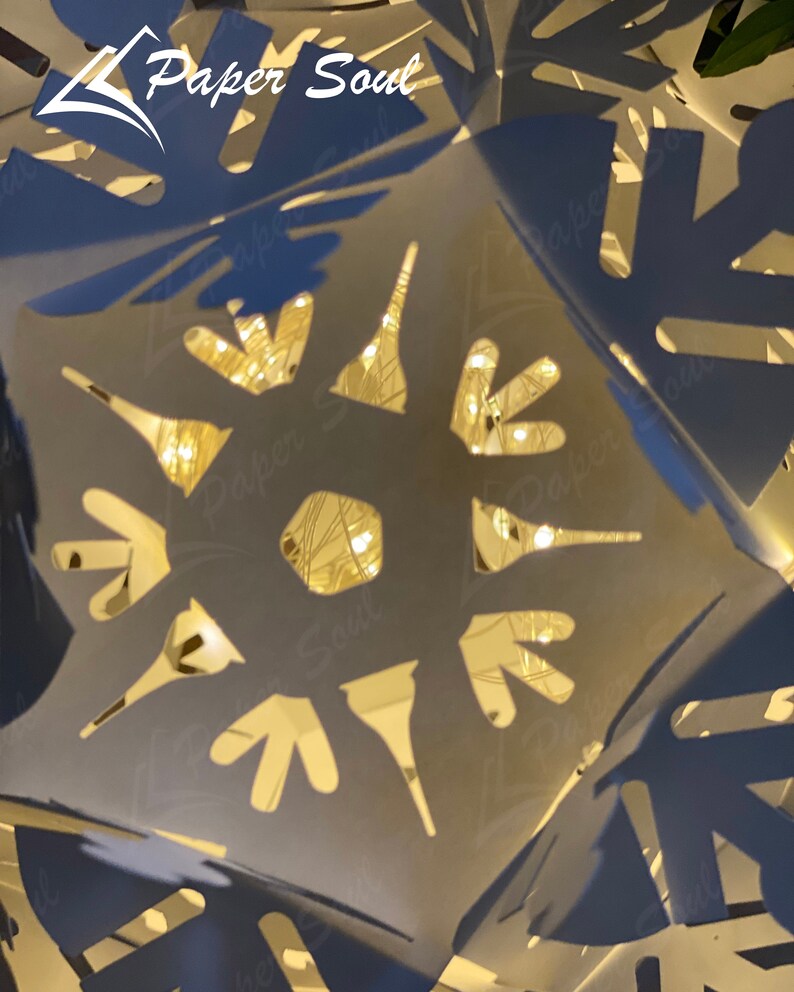 Peut inclure: Une flocon de neige en papier blanc avec des d&eacute;coupes complexes, &eacute;clair&eacute; par des lumi&egrave;res blanches chaudes. Le flocon de neige est pli&eacute; en une forme tridimensionnelle, cr&eacute;ant un design d&eacute;licat et festif. Le texte "Paper Soul" est visible dans le coin sup&eacute;rieur gauche de l'image.