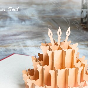 以下が含まれることがあります： 3本のろうそくが立てられた誕生日ケーキが描かれた3Dポップアップカード。ケーキは紙製で、薄い茶色です。カードは開いており、ケーキは立っています。背景は木製の表面のぼやけた画像です。