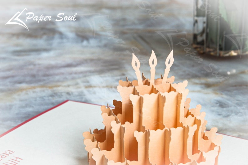 以下が含まれることがあります： 3本のろうそくが立てられた誕生日ケーキが描かれた3Dポップアップカード。ケーキは紙製で、薄い茶色です。カードは開いており、ケーキは立っています。背景は木製の表面のぼやけた画像です。