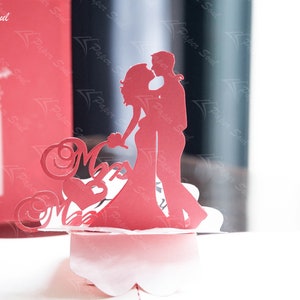 Pode incluir: Um cart&atilde;o pop-up de papel vermelho com uma silhueta de um casal dan&ccedil;ando. O cart&atilde;o &eacute; decorado com recortes de papel intrincados e as palavras "Mr & Mrs".