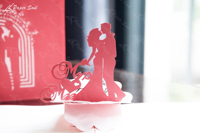 Pode incluir: Um cart&atilde;o pop-up de papel vermelho com uma silhueta de um casal dan&ccedil;ando. O cart&atilde;o &eacute; decorado com recortes de papel intrincados e as palavras "Mr & Mrs".