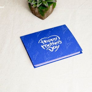 May include: A blue card with a white heart that says "Happy Mother's Day" in a handwritten font. The card has a subtle pattern of the words "Paper Soul" repeated on it.