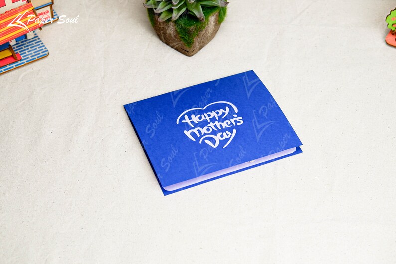 May include: A blue card with a white heart that says "Happy Mother's Day" in a handwritten font. The card has a subtle pattern of the words "Paper Soul" repeated on it.