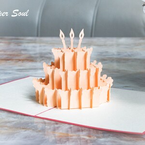 以下が含まれることがあります： 3本のろうそくが立てられた誕生日ケーキが描かれた、桃色の3Dポップアップカード。カードが開いており、ケーキはカードの中央にあります。カードは木製のテーブルの上に置かれています。