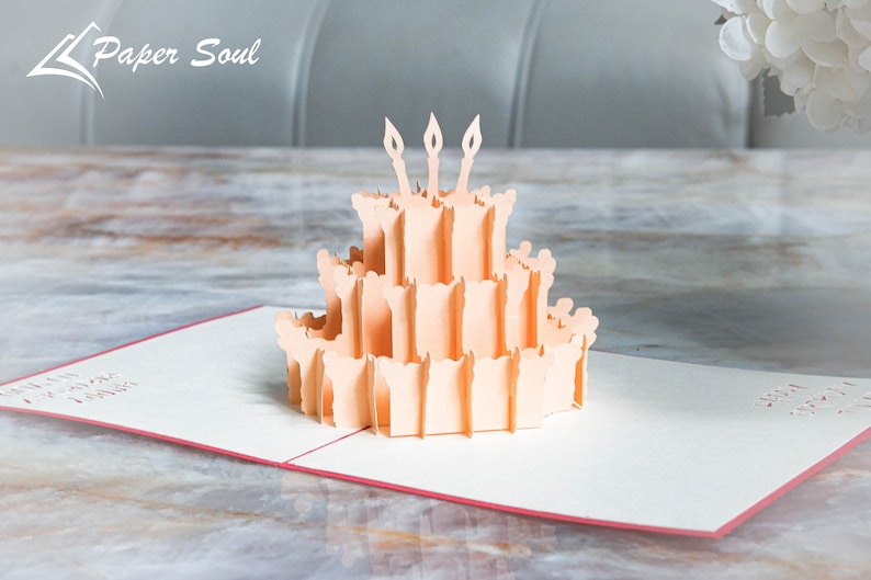 以下が含まれることがあります： 3本のろうそくが立てられた誕生日ケーキが描かれた、桃色の3Dポップアップカード。カードが開いており、ケーキはカードの中央にあります。カードは木製のテーブルの上に置かれています。