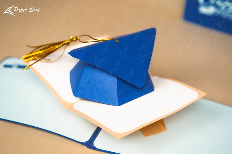 May include: A blue graduation cap shaped box with a gold tassel. The box is open and sitting on a white card with a brown border. The card is on top of a blue surface.