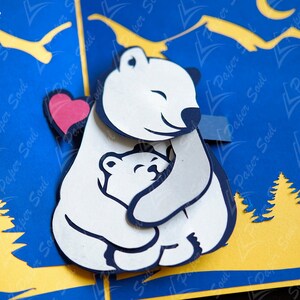 May include: A white papercraft polar bear with a red heart cutout, hugging a smaller white polar bear. The bears are set against a blue background with yellow mountains and trees.