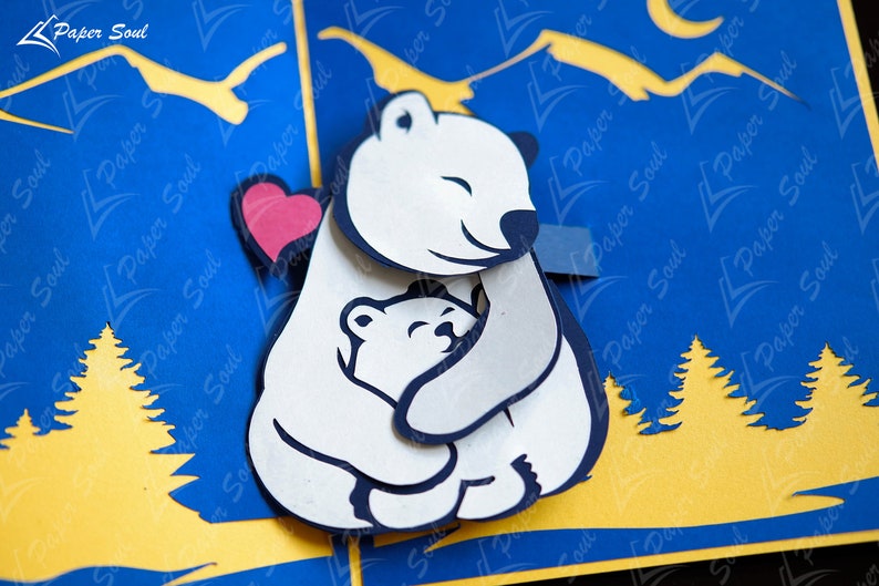 May include: A white papercraft polar bear with a red heart cutout, hugging a smaller white polar bear. The bears are set against a blue background with yellow mountains and trees.