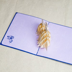 May include: A light purple pop-up card with a blue border. The card is open and features a 3D paper sculpture of two people holding hands. The card has a blue rose design on the front.