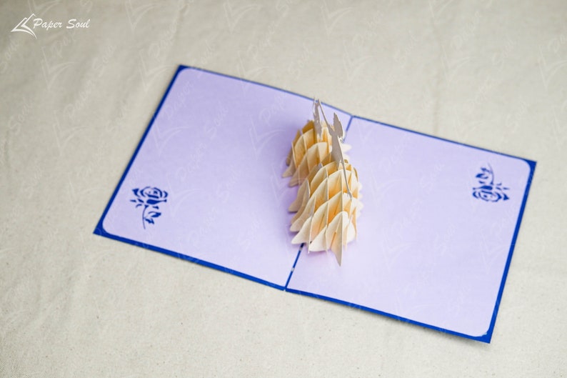 May include: A light purple pop-up card with a blue border. The card is open and features a 3D paper sculpture of two people holding hands. The card has a blue rose design on the front.