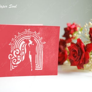 Pode incluir: Cart&atilde;o de papel recortado vermelho com uma silhueta de um casal em p&eacute; sob um arco decorado com flores e espirais. O cart&atilde;o est&aacute; sobre uma superf&iacute;cie branca com rosas vermelhas e flores brancas no fundo.