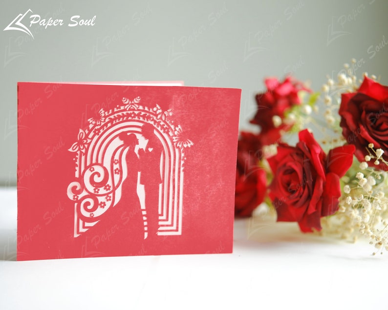 Pode incluir: Cart&atilde;o de papel recortado vermelho com uma silhueta de um casal em p&eacute; sob um arco decorado com flores e espirais. O cart&atilde;o est&aacute; sobre uma superf&iacute;cie branca com rosas vermelhas e flores brancas no fundo.