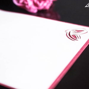 Pode incluir: Um cart&atilde;o branco com uma borda rosa e um design recortado de dois an&eacute;is entrela&ccedil;ados. O cart&atilde;o est&aacute; sobre uma superf&iacute;cie preta.