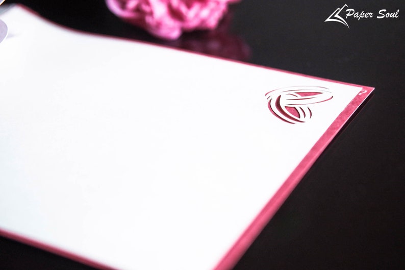 Pode incluir: Um cart&atilde;o branco com uma borda rosa e um design recortado de dois an&eacute;is entrela&ccedil;ados. O cart&atilde;o est&aacute; sobre uma superf&iacute;cie preta.