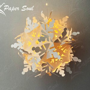 Peut inclure: Une d&eacute;coration de No&euml;l en forme de flocon de neige en papier blanc avec une lumi&egrave;re blanche chaude qui brille &agrave; travers les d&eacute;coupes complexes. Le flocon de neige est suspendu au plafond et le texte "Paper Soul" est visible dans le coin sup&eacute;rieur gauche de l'image.