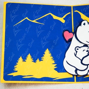May include: A white polar bear with a red heart cutout holding a smaller white polar bear. The bears are against a blue background with yellow mountains and trees.