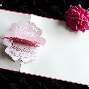 Pode incluir: Um cart&atilde;o pop-up rosa e branco com as palavras "Happy Wedding" em uma fonte decorativa. O cart&atilde;o est&aacute; aberto e o pop-up tem a forma de uma flor. Uma rosa rosa est&aacute; no fundo.