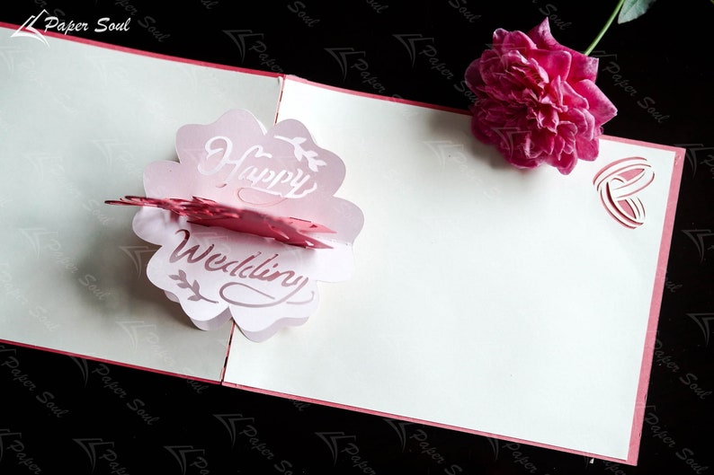 Pode incluir: Um cart&atilde;o pop-up rosa e branco com as palavras "Happy Wedding" em uma fonte decorativa. O cart&atilde;o est&aacute; aberto e o pop-up tem a forma de uma flor. Uma rosa rosa est&aacute; no fundo.