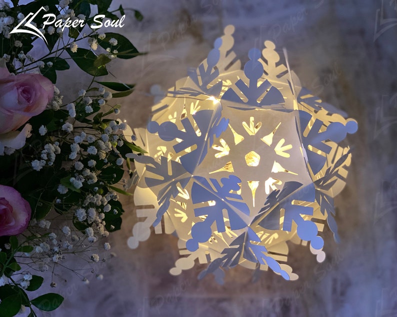 Peut inclure: Une lanterne en papier en forme de flocon de neige blanche avec une lumi&egrave;re &agrave; l'int&eacute;rieur. La lanterne est faite de plusieurs couches de papier et pr&eacute;sente un design d&eacute;licat et complexe. La lumi&egrave;re traverse le papier, cr&eacute;ant une lueur chaleureuse et accueillante.