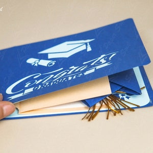 May include: A blue pop-up card with a graduation cap and the word "Congrats!" on the front. The card is open to reveal a layered design with a gold tassel.