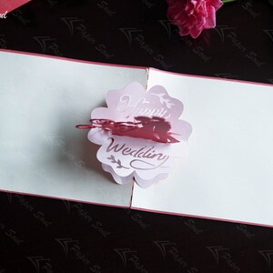 Pode incluir: Um cart&atilde;o pop-up 3D rosa e branco com um design de flor e as palavras "Happy Wedding" no interior. O cart&atilde;o est&aacute; fechado e tem uma borda vermelha.
