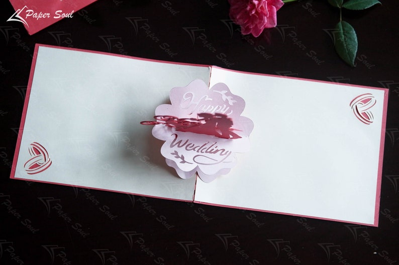 Pode incluir: Um cart&atilde;o pop-up 3D rosa e branco com um design de flor e as palavras "Happy Wedding" no interior. O cart&atilde;o est&aacute; fechado e tem uma borda vermelha.