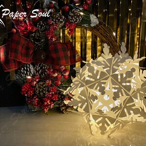 Peut inclure: Une d&eacute;coration de flocon de neige en papier blanc avec une lumi&egrave;re &agrave; l'int&eacute;rieur. Le flocon de neige est &eacute;clair&eacute; et diffuse une lueur chaude. Le flocon de neige est pos&eacute; sur une table devant une couronne de No&euml;l avec des baies rouges et un n&oelig;ud &agrave; carreaux.
