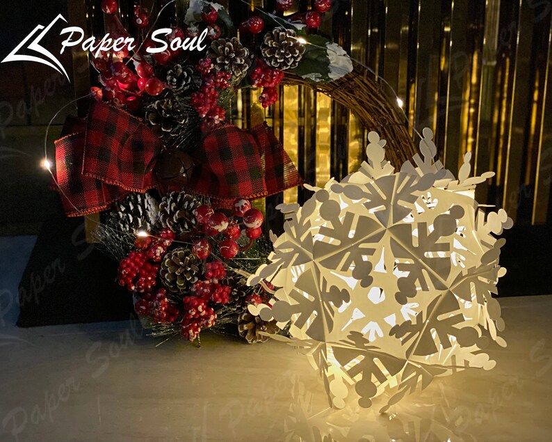 Peut inclure: Une d&eacute;coration de flocon de neige en papier blanc avec une lumi&egrave;re &agrave; l'int&eacute;rieur. Le flocon de neige est &eacute;clair&eacute; et diffuse une lueur chaude. Le flocon de neige est pos&eacute; sur une table devant une couronne de No&euml;l avec des baies rouges et un n&oelig;ud &agrave; carreaux.