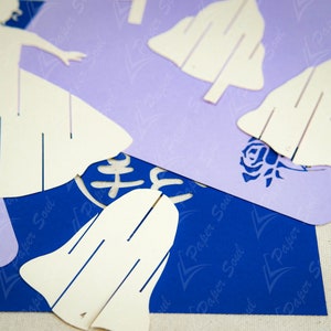 May include: White paper cutouts of a woman in a dress and several bells. The cutouts are on top of a blue and purple paper background with the text "Paper Soul" repeated on the background.