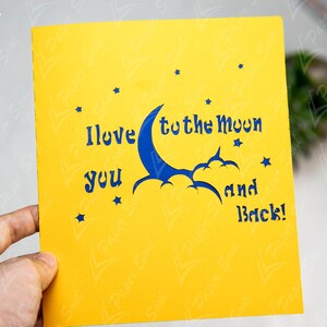 May include: A yellow card with a blue crescent moon and stars cut out. The card reads "I love you to the moon and back!"