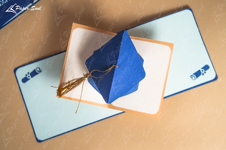 May include: A blue graduation cap shaped box with a gold tassel, sitting on a blue and white card with a diploma icon. The card is on a brown surface.
