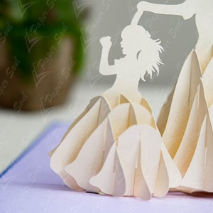 May include: A white paper pop-up card with a 3D design of a girl dancing. The girl is holding up her arms and has a long flowing skirt. The card is made of intricate paper cuts and is a beautiful example of paper art.