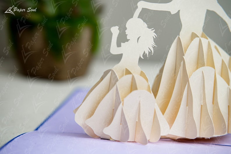 May include: A white paper pop-up card with a 3D design of a girl dancing. The girl is holding up her arms and has a long flowing skirt. The card is made of intricate paper cuts and is a beautiful example of paper art.