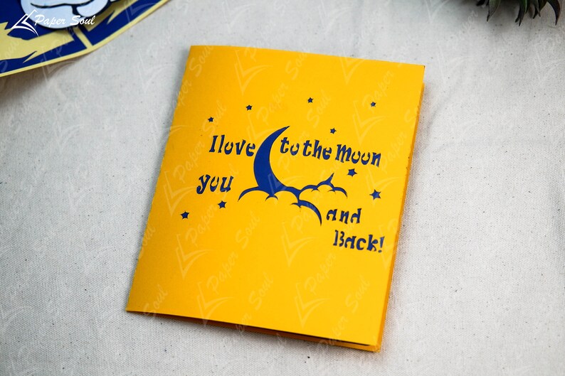May include: A yellow card with a blue crescent moon and clouds, and the text "I love you to the moon and back!" with stars around the moon.