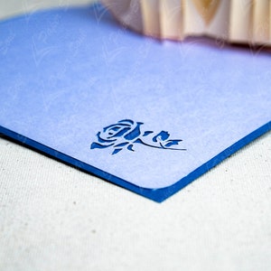 May include: A light blue card with a blue border and a cut-out design of a rose. The card is on a white textured surface.