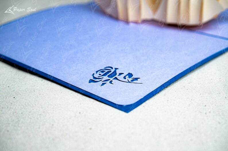 May include: A light blue card with a blue border and a cut-out design of a rose. The card is on a white textured surface.