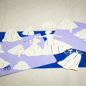 May include: White paper cutouts of two figures in dresses, a larger one holding the hand of a smaller one, and several paper cutouts of bell shapes on a purple and blue background.