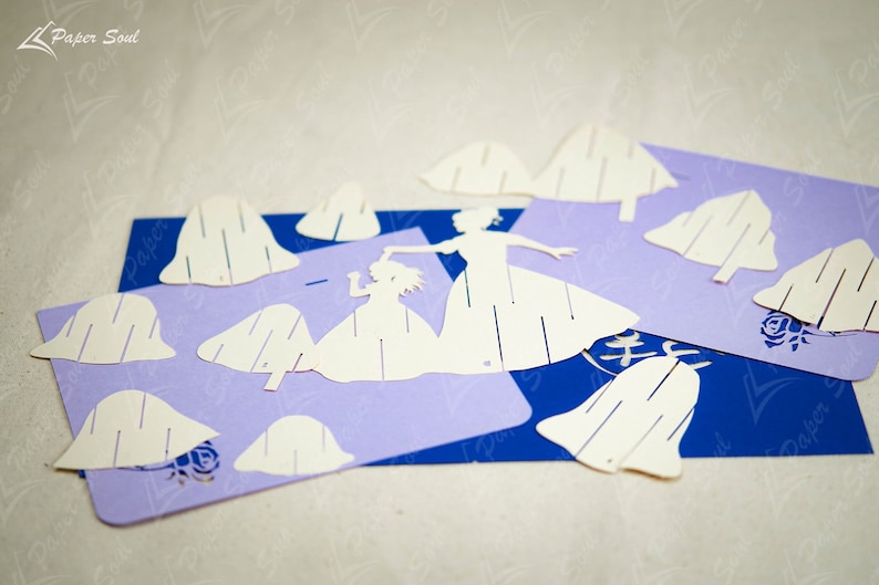 May include: White paper cutouts of two figures in dresses, a larger one holding the hand of a smaller one, and several paper cutouts of bell shapes on a purple and blue background.